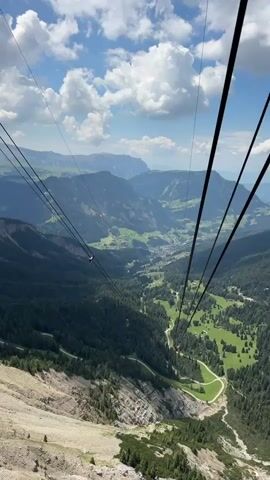 The video shows a stunning aerial view of a valley from a cable car. The cable car cables are prominently visible in the foreground, stretching diagonally across the frame. Below, a lush green valley unfolds, dotted with scattered trees and winding paths. In the distance, majestic mountains rise, their slopes covered in dense forests. The sky is a vibrant blue, adorned with fluffy white clouds, suggesting a pleasant, possibly midday, weather condition. The scene evokes a sense of adventure and awe, as the viewer is transported over the breathtaking mountain landscape. The presence of a few small red dots in the distance, possibly other cable cars or gliders, adds a dynamic element to the scenery. The location is identified as Urtijëi - St. Ulrich in Gröden - Ortisei, Italy, a region known for its spectacular Dolomite scenery and popular for outdoor activities. The overall mood is one of exhilaration and natural beauty. No text is visible in the video.