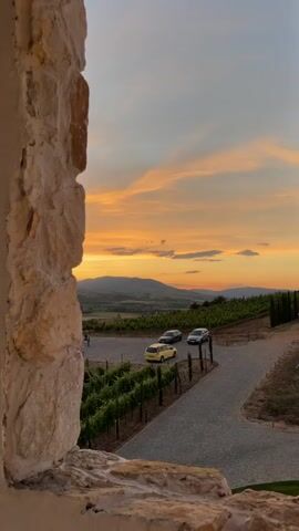 The video opens with a close-up of a rough, stone-encrusted wall, partially obscuring the view beyond. The camera pans slowly to the right, revealing a rustic archway framing a picturesque landscape. A vineyard stretches out below, with neat rows of green vines leading towards rolling hills bathed in the warm glow of a sunset. Several cars are parked on a paved path winding through the vineyard. The sky above is a canvas of orange, pink, and yellow, with wispy clouds adding texture to the scene. As the camera continues to move, more arched openings appear, each offering a slightly different perspective of the same serene vista. The video ends with a view of two large bells hanging from the ceiling, silhouetted against the twilight sky.