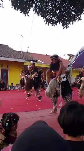The video shows an outdoor performance on a red carpeted stage. Several performers, dressed in traditional attire including headdresses and loincloths, are dancing. Some of the performers are holding and manipulating horse-like props made of white horsehair, which they appear to be riding or embodying. In the foreground, a group of spectators, mostly children and adults with their backs to the camera, are watching the performance. Behind the stage, there are buildings with yellow walls and tiled roofs, and trees overhead. A purple and white tent structure is visible behind the performers, serving as a backdrop or canopy. A banner with text is partially visible on the stage. The lighting suggests it is daytime.