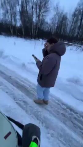 The video shows a person standing on a snow-covered road while using a smartphone. The person is wearing a hooded jacket, light-colored pants, and tan boots. The road is icy and has tire tracks. There is a snow-covered field on either side of the road, with bare trees in the background. The sky is overcast. The camera appears to be attached to or held by a person pushing a stroller.