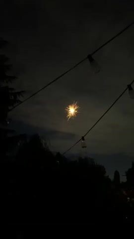 The video shows a nighttime scene with fireworks exploding in the sky. String lights with exposed bulbs are visible in the foreground, strung across the frame. Silhouettes of trees and foliage are visible at the bottom of the frame, suggesting an outdoor setting. The fireworks are primarily yellow and red, with a few bursts of white light. The sky is dark, with some clouds visible. There are no people clearly visible in the frame.