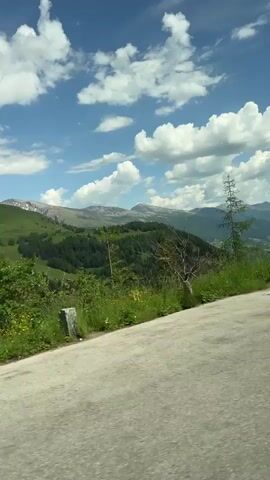 The video shows a scenic view of mountains and green hills. The sky is blue with fluffy white clouds. There are pine trees and other vegetation visible. The camera is moving, suggesting it is being filmed from a vehicle. There are no people visible in the video.