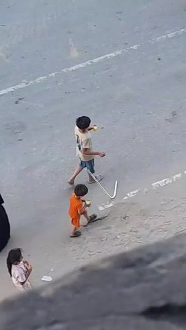 Two young boys are walking away from the camera. The boy in front is wearing an orange shirt and dark shorts. He is carrying a white object in his right hand and a yellow cup in his left hand. The boy behind him is wearing a tan t-shirt with a graphic design on the back and blue shorts. He is also carrying a yellow cup in his right hand. The ground is paved and appears to be a street or walkway. There is a white line painted on the ground. The boys are walking towards the right side of the frame.