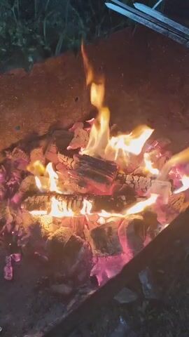 A close-up shot focuses on a campfire. Flames dance and flicker over burning logs and glowing embers. The fire is contained within a dark, metal fire pit. The background is dark and out of focus, suggesting the scene is taking place outdoors at night. The overall impression is one of warmth and natural light.