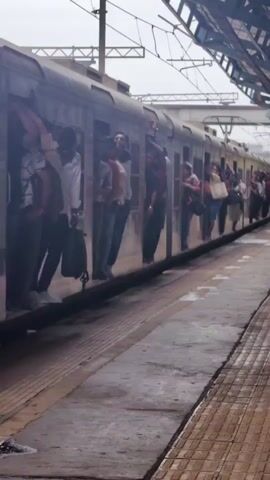 A crowded train is stopped at a station platform. Several people are hanging out of the train doors and windows. The train is silver and appears to be an commuter train. The platform is paved with checkered tiles and there are overhead wires and a canopy visible. The sky is overcast.