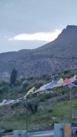 This media file captures a scenic view of a mountainous landscape in Kead, India. Prayer flags, in various colors including blue, white, red, green, and yellow, are strung across the foreground, gently fluttering in the breeze. They are attached to metal poles, with one pole visible on the right side of the frame.

Behind the prayer flags, a lush green hillside is dotted with sparse vegetation and small white wildflowers. A solitary dark green tree stands on the left side of the hill. The midground is dominated by a rugged, dark-colored mountain that rises majestically against the sky. The mountain's textured surface suggests it is covered in small shrubs or rocks.

The sky above is a blend of blue and grey, with a prominent band of white clouds illuminated by sunlight, suggesting it is either sunrise or sunset. The lighting conditions create a somewhat dramatic and serene atmosphere. The overall scene evokes a sense of peace and spirituality, characteristic of mountainous regions often associated with Buddhist traditions where prayer flags are commonly used. The terrain appears dry and arid, typical of high-altitude areas. There is no visible text in the media file.