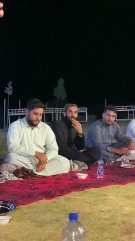 The video shows a group of men sitting on a red patterned rug on the grass at night. The men are dressed in traditional South Asian attire. They appear to be having a meal, with plates of food and bottles of water visible on the rug. One man in the center is speaking with his hand near his mouth. Another man to his right is looking down at the food. A third man to his left is looking forward and appears to be listening. In the background, there are white fences and some trees. The lighting is dim, suggesting it is evening or nighttime.
