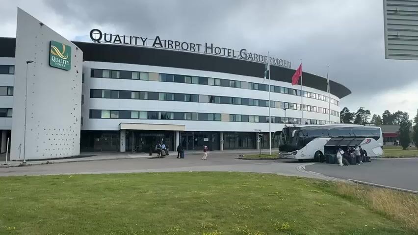 The video shows the Quality Airport Hotel Gardermoen. A bus is parked in front of the hotel, and several people are standing next to it with luggage. Some people are entering and exiting the hotel's main entrance. The sky is cloudy.