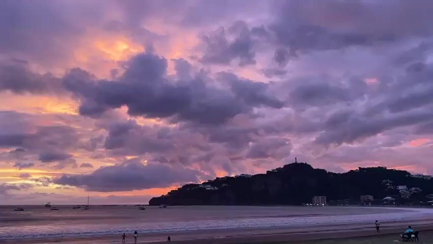 The video shows a wide shot of a beach with a dramatic sunset. The sky is filled with colorful clouds, ranging from pink and orange to deep purple. A dark, hilly peninsula with buildings and a statue on top juts out into the ocean on the right side of the frame. Several sailboats are anchored in the water near the peninsula. The ocean has gentle waves, and the beach in the foreground is sandy. A few people are visible on the beach, some walking and others sitting. The overall atmosphere is serene and picturesque.