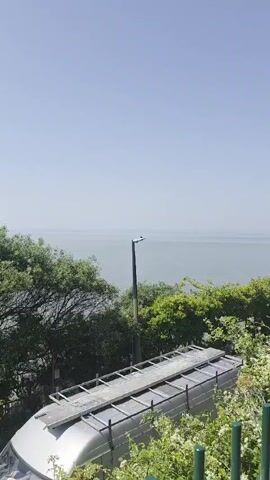 This video captures a scenic view of the sea and coastline at Weston-super-Mare, United Kingdom. The scene is bathed in bright sunlight, indicating a clear and sunny day, likely late morning or early afternoon given the quality of light.

The primary subjects are the expansive sea and sky, which dominate the upper portion of the frame. In the foreground, several objects are visible. There's a section of what appears to be a roof or platform, possibly from a building or vehicle, with metal framing. To the right, a tall lamppost stands against the backdrop of the sea.

Further out, a pier stretches into the water. There's evidence of construction or renovation work on or around the pier, with a large crane positioned nearby and scaffolding visible. A small island or distant landmass can be seen on the horizon, adding depth to the seascape.

Green trees and foliage are scattered throughout the scene, particularly in the middle ground, softening the man-made structures and the industrial elements like the crane. A van with a roof rack is parked in the lower right section of the frame, suggesting the presence of human activity.

The overall mood is one of a bright, possibly coastal industrial or recreational area on a pleasant day. There are no people clearly visible in the video, and the focus remains on the landscape and the structures within it. The activity appears to be ongoing construction at the pier, contrasted with the tranquility of the sea and sky.