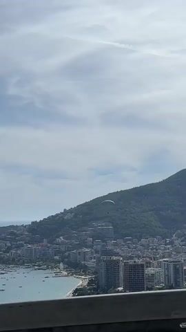 A paraglider is visible in the sky above a green, tree-covered mountain. Below the mountain is a city with buildings of varying heights, and a body of water with boats. The sky is partly cloudy. The video appears to be filmed from a moving vehicle, as indicated by the blurry foreground element which could be a guardrail.