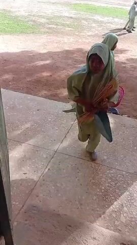 A girl in a green hijab and matching pants sweeps the ground with a broom. She is wearing white shoes. Another girl in a similar green outfit stands in the background holding a pink basket filled with what appear to be trash items. The ground is made of concrete with a rough texture, and there is dirt and sand visible. The scene appears to be outdoors, possibly in a schoolyard or public space, with trees and shadows casting across the ground. The camera is positioned at an angle, looking down at the girls and the area they are in.