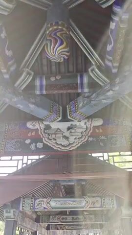 This is a video of the interior of a covered walkway with a wooden roof and columns. The structure is ornately decorated with colorful paintings and carvings in a traditional East Asian style. The camera pans slowly upwards, showing the intricate details of the ceiling beams and decorative elements, including what appears to be a colorful spiral ornament at the very top. Below the ceiling, the walkway extends into the distance, with more columns and decorative panels visible. Sunlight streams in from the sides, casting shadows on the walkway. A person in the foreground is holding a smartphone and appears to be taking a picture or video. Another person is visible further down the walkway. Lush green foliage is visible through the open sides of the walkway.