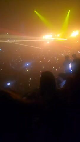 The video shows a large crowd at a concert venue, from a high vantage point. The audience holds up phones, with their screens illuminating the scene. A stage is visible, featuring a performer on a platform. Stage lighting creates a smoky atmosphere, with beams of yellow and red light. The backdrop behind the performer cycles through different colors and patterns. The camera appears to be held by an audience member.