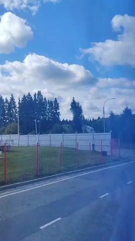 A white truck is seen driving past a metal fence on a sunny day. The sky is blue with white clouds. There are trees and a house in the background.