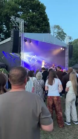 The video shows an outdoor concert. In the foreground, several people are standing, with the back of a man's head and shoulders visible in the lower left. Behind them, a crowd watches a band performing on a stage. The stage is set up with lighting, speakers, and a canopy. A female singer is at the front of the stage, and other band members, including a drummer, are visible behind her. The concert appears to be taking place in the evening, with the sky a clear blue. Trees and some tents are visible in the background. The overall atmosphere seems relaxed and festive.
