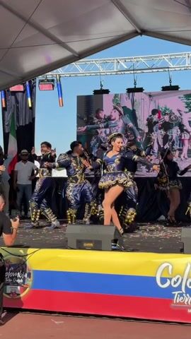 The video shows a dance performance on a stage under a tent. There are several dancers in ornate costumes, some with tall hats and embellished jackets and pants, and others in shorter, ruffled skirts. The dancers are moving in synchronized steps, some in groups and others individually. A large screen behind the performers displays images, possibly of previous performances or related content. In the foreground, there's a banner with the Colombian flag colors and the words "Colombia Tierra Querida" and "Milan-Italia". Confetti is scattered on the stage. There are speakers visible on the stage floor. In the background, outside the tent, a blue sky and some foliage can be seen, along with some people standing and observing. The overall atmosphere appears to be festive, suggesting a cultural event or festival.