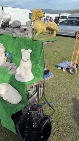 The video shows a display of garden ornaments set up in a grassy area, likely at an outdoor market or fair. The display is arranged on a table covered with artificial turf. A variety of concrete animal figures are visible, including several dog statues, one of which is a water feature with water trickling into a black bucket below. A blue and yellow parrot statue and a stone face are also part of the display. In the background, there are parked cars, vans, and people milling about. A hand truck and a dolly are visible near the display. The sky is overcast, suggesting a cloudy day. The scene is busy with activity, typical of a market or fair environment.