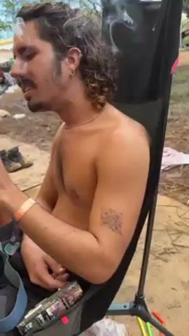 A man with long curly hair and a mustache sits shirtless in a black camping chair outdoors. He has a geometric tattoo on his left upper arm and wears a silver chain necklace and a wristband. He is smoking, and there is a can of drink in the chair beside him. The ground is muddy and littered with straws. People and tents are visible in the background.