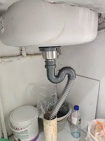 The video shows the underside of a sink with its plumbing. A gray P-trap is connected to the sink drain, and a flexible gray corrugated pipe extends from the P-trap downwards into a vertical pipe. To the left of the plumbing, there is a white bucket with a red and blue label and a plastic lid. To the right of the plumbing, there is a bucket containing some dark material, a clear plastic bottle with a blue cap, and another white bucket. The background consists of white walls and some tile work above the sink. The overall environment appears to be utilitarian, possibly a utility area or under a kitchen sink.