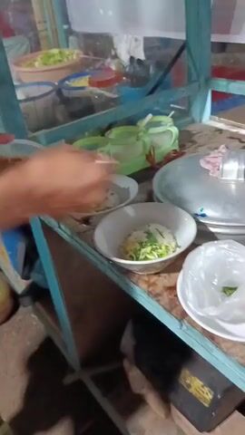 The video shows a close-up of a food stall, likely preparing to serve customers. The camera focuses on a wooden cart with a blue painted frame and a clear plastic barrier at the top. On the cart, there are several bowls of food, appearing to be noodles or soup with vegetables. There are also small condiment containers and a large metal pot with a lid. To the left, a person's hand is visible holding a jar of red sauce. The lighting suggests it is evening or nighttime.