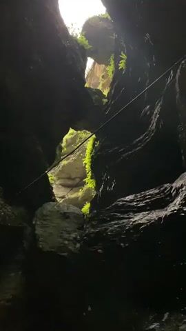 This is a video taken from within a narrow canyon, looking upwards towards the sky. Dark, sheer rock walls rise on either side, with patches of lush green vegetation clinging to them. A thin, dark rope or cable is stretched diagonally across the frame, disappearing into the shadows. Light filters in from the opening above, illuminating the upper portions of the canyon walls and a natural archway formed by the rock. The bottom of the canyon appears dark and rocky. The camera is angled upwards, creating a sense of depth and the imposing scale of the natural formation.