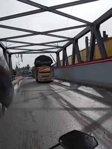 The video shows a convoy of trucks on a road that appears to be a bridge. The first truck is yellow and is covered with a black tarp. Behind it, there are several other trucks, some of which are also yellow and covered. Two motorcycles are visible, one carrying two people and another carrying a single person. The road surface is wet and appears to be paved. The sky is overcast. The scene suggests a traffic jam or a slow-moving convoy.