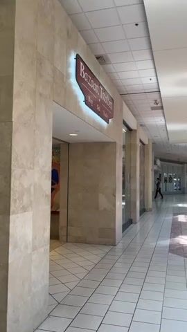 The video shows a shopping mall corridor with a store called Bazaar Trader. The store has a sign that says "Bazaar Trader" and "Buy, Sell, Trade." The store's interior is visible, showing shelves filled with various items. The corridor has tiled floors and a dropped ceiling with fluorescent lighting. Sunlight streams in from skylights in the ceiling, creating shadows on the floor. The mall appears to be quiet, with no other people visible in the immediate vicinity.