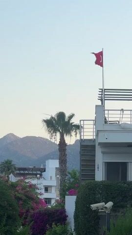This vertical, outdoor video captures a close-up view of a building with a Turkish flag flying from its rooftop. The flag, red with a white star and crescent, is prominent against a pale sky. In the midground, a tall palm tree stands to the left of the flag pole. Behind the palm tree and the building, a range of rugged, mountainous terrain stretches across the horizon.

The building itself is modern, with white walls and a balcony area featuring white railings. There are also two security cameras visible at the bottom right of the frame, mounted on a dark post. Lush greenery, including vibrant purple bougainvillea, surrounds the base of the building, adding a touch of color to the scene. Several other buildings, also appearing modern and white, are visible in the background, interspersed with more greenery.

The scene appears to be set during the late afternoon or early evening, suggested by the soft, diffused light and the clear sky, which lacks the harsh shadows of midday sun. The weather seems calm and pleasant, with no visible wind disturbing the foliage excessively, though the flag is gently rippling. The overall impression is one of a peaceful, sunny day in a coastal or hilly region, likely a residential or tourist area, given the style of architecture and the presence of security cameras. The location is identified as Marmaris, Turkey, which aligns with the visual cues of the palm tree, mountainous backdrop, and the Turkish flag. There is no discernible text within the video frame.