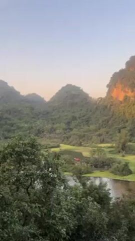 The video shows a panoramic view of a tropical landscape with lush green hills, a body of water, and clear sky. The hills are covered with dense vegetation, and the water reflects the sky. A red boat is visible on the water. The lighting suggests it is either early morning or late afternoon.