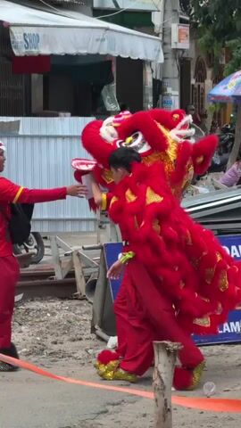 Two individuals are performing a dragon dance. The person in front is wearing a red dragon costume with gold accents. The person behind is also in a red costume, holding a red tray. They are on a street with buildings and parked motorcycles in the background.