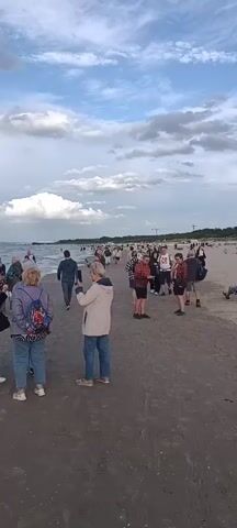 The video shows a beach with many people. The sky is cloudy with patches of blue. There are waves crashing on the shore. Several people are walking along the beach, some are standing and talking, and one person is sitting on a chair in the distance. The sand is dark and wet near the water. There are some plants and dunes in the background. The overall atmosphere appears to be casual and leisurely.