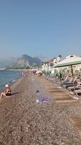 The video shows a pebble beach with several people. On the left, a woman in a pink hat and black swimsuit sits on the shore. In the water, several people are swimming and wading. A man in blue shorts walks out of the water. In the background, there are mountains and a clear blue sky. On the right side of the beach, there are rows of empty lounge chairs under umbrellas.