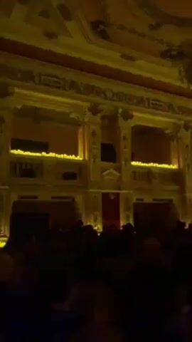 The video shows the interior of a theater with a crowd of people in the foreground, facing a stage. The stage has a red curtain. Above the stage, there are ornate balconies and columns, lit with yellow light. The ceiling is decorated with geometric patterns and a chandelier. The overall lighting in the theater is dim. Onstage, figures can be seen faintly. The camera slowly pans across the room.