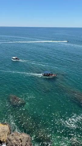 The video shows a view from a cliff overlooking a bright blue ocean on a clear day. The water is a vibrant turquoise color, with sunlight glinting off the surface. Two boats are visible in the water. The closer boat is a small, open vessel with a dark blue canopy, moving away from the viewer towards the left of the frame. It appears to have several people on board. Further out in the distance, another smaller boat is visible, also moving away from the viewer towards the left. There are some dark rocks partially submerged in the turquoise water near the cliff edge. The horizon is clear and meets the bright blue sky.