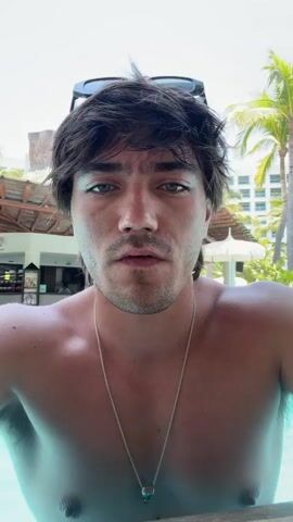 A person with brown hair, hazel eyes, and a small beard is sitting shirtless in a pool, outdoors. Sunglasses are on top of his head, and a silver necklace hangs around his neck. He’s holding a green smoothie with an orange slice on the rim and a straw in the cup. Palm trees and a building are visible in the background. The person says, “Girl's breakfast,” and then drinks the smoothie through the straw.