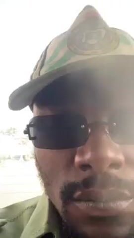 The video shows a man wearing sunglasses and a green hat with an emblem, possibly military-related. He is likely inside a vehicle, looking out of the back window. Through the dusty window, a street scene is visible with buildings, vehicles, and palm trees under a hazy sky. The video is shaky, suggesting movement of the vehicle.