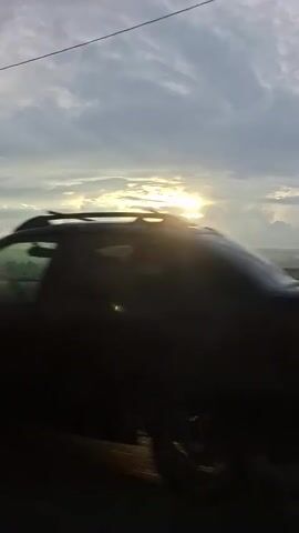 A car is seen driving on a wet road. The sun is visible in the sky, peeking through the clouds. The landscape is hazy and appears to be rural, with houses and trees visible in the distance. The overall mood is peaceful and serene.