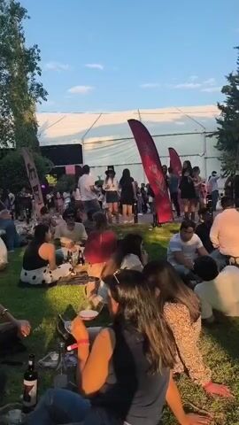 The video shows a large group of people gathered on a grassy area, likely at an outdoor event or festival. Many are sitting on the grass in small groups, seemingly enjoying picnics with food and drinks. There are bottles of wine and glasses visible. The atmosphere appears relaxed and social. In the background, there is a large white tent-like structure. Red banners are scattered throughout the area. A man dressed in black walks through the crowd, holding a bottle in his hand. The weather appears to be sunny.