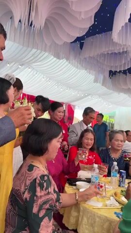 This video captures a lively outdoor celebration, likely a wedding, in Tay Son Commune, Vietnam. The scene is set under a large white tent adorned with elegant white fabric drapes and string lights, with some red and blue accents visible in the background.

Numerous people, predominantly adults, are present. Guests are seated at round tables covered with yellow tablecloths, engaged in conversation, eating, and drinking. Many attendees are smiling, conveying a cheerful and celebratory atmosphere. A bride, wearing a white wedding gown and veil, is seen interacting with guests at a table, holding a white floral bouquet. Several women are dressed in vibrant traditional Vietnamese attire, including red and pink Ao Dai-style tops.

The tables are laden with food and drinks. Visible dishes include what appears to be grilled or fried chicken, fresh herbs, and other local delicacies, served on large round trays. Beverages include bottled water (with "NƯỚC UỐNG TINH KHIẾT" and "TRÀ ĐÁ" labels partially visible) and a can of "HA NOI BEER." Guests are using chopsticks to eat. A pink, compact gimbal camera mounted on a small tripod is also seen on one of the tables, suggesting that the event is being recorded. The lighting suggests it is daytime.