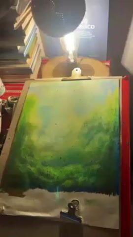 The video shows a person holding a decorative gourd lamp with holes that create a pattern when lit. The lamp casts light onto a painting in progress. The painting is a landscape with green and blue hues. It is clipped to a board that sits on a red table. In the background, there are books and other items.