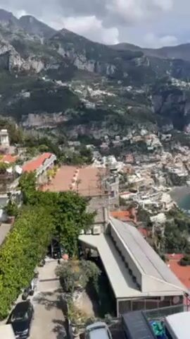The video shows a coastal town nestled on a steep hillside, with the sea visible in the background. The town is made up of closely packed white buildings with orange tile roofs. Lush green vegetation covers the hillside and surrounding mountains, which rise dramatically in the background. The sea is a deep turquoise color. In the foreground, there are buildings with visible rooftops and parked cars. The sky is partly cloudy.
