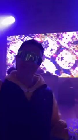 A person wearing a black vest, yellow jacket, and neon green sunglasses is holding a cup and dancing in a room lit with blue light.  In the background is a large screen with a chaotic image of red, purple, and white shapes. Further back is a DJ booth and a person standing behind it, silhouetted by the light from the screen.  The person in the foreground raises their arm and waves their hand as they dance.