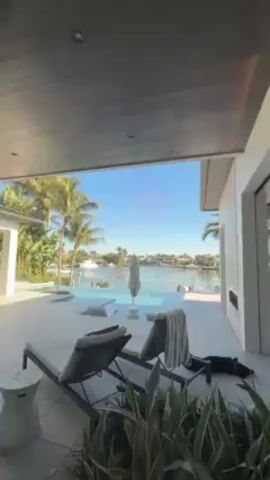 The video shows the interior of a modern house with a view of a waterway through a glass sliding door. The camera pans from a patio area with lounge chairs to an open-plan dining and kitchen area. The dining area has an oval table set for several people, with chairs upholstered in a floral pattern. The kitchen features white cabinets, stainless steel appliances, and a central island. A person dressed in a white shirt and black pants is bending down near a cabinet on the right side of the kitchen. A glass-fronted cabinet is visible behind the person, displaying various items. The lighting is modern, with recessed ceiling lights and a decorative light fixture above the dining table. The video focuses on showcasing the interior design and layout of the house.