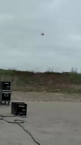 The scene shows a helicopter flying against a cloudy sky, with a fire effect shooting flames upward from a black device situated on a concrete surface. Adjacent to the device on its left are two black cases stacked on top of each other, also resting on the concrete. The ground is barren, with a grassy embankment in the background. Electrical cords connect the devices.