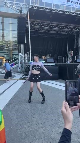 A person with dark, short hair, wearing a black crop top with a mesh overlay, a black mini skirt with butterfly accents, and black platform boots with buckles, walks in front of a stage at an outdoor event. The person has on colorful butterfly-shaped accessories on their shoulders. They are holding a fan in their right hand. To the right of the person, a person wearing a black shirt and headphones is filming with a smartphone. Further to the right, another person is partially visible from behind, also seemingly observing. To the left of the person, a person in a blue t-shirt and black shorts stands near some equipment. Behind the person on stage, a black curtain covers the stage, with drums and speakers visible. Above the stage, a metal truss structure supports lighting and speakers, and a banner with "SURREY PRIDE.CA" is visible. A building with large windows is seen in the background. The ground is paved with interlocking bricks.