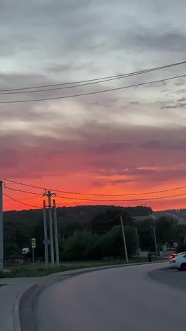 The video shows a vibrant sunset over a rural landscape. The sky is a mix of deep red and shades of gray. Utility poles and power lines cross the scene. A road curves into the frame, with a sidewalk and grass on the left. Some trees, possibly part of a forest, are visible in the distance. A few buildings and a white car are parked along the road. Two people walk along the right side of the street. There are pedestrian crossing signs visible.