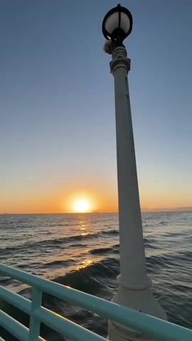 The video is a vertical, handheld shot of a pier railing and lamppost against a sunset over the ocean. The sun is a bright orb on the horizon, casting a golden glow on the water. Gentle waves lap against the pier. The sky is a clear, pale blue, transitioning to orange and pink near the horizon. The lamppost is tall and ornate, with a black lantern at the top. The railing is painted a light teal color. The video is approximately 4 seconds long and appears to have been filmed in natural light during sunset.