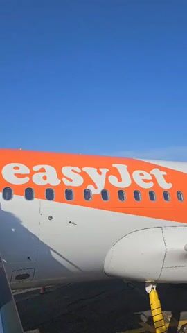 This is a vertical video taken from the perspective of an airplane passenger. The video begins with a close-up of the side of an easyJet airplane, showing the orange and white livery and several windows. The camera then pans to the right, revealing the rear of the plane with the tail fin prominently displaying the easyJet logo. A set of stairs, labeled "CASAVIA," leads up to the plane's entrance, and a group of passengers, carrying luggage, are seen boarding. In the background, a truck is positioned near the stairs. Beyond the aircraft and the tarmac, a mountainous landscape with a clear blue sky can be observed. The video continues to show more passengers on the stairs and the ground, with the airplane and the mountainous backdrop remaining visible.