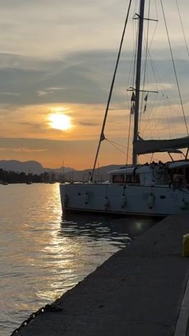 The video captures a serene sunset scene at a harbor in Poros, Greece. A large white catamaran, featuring a tall mast and rigging, is moored alongside a concrete dock on the right side of the frame. The boat has several white buoys hanging from its side, and the text "400 S2" is visible on its stern.

The sun is low in the sky, positioned centrally, casting a warm golden glow that reflects brightly on the calm water surface. The sky is a mix of orange and light blue, with scattered clouds. In the background, across the water, silhouettes of mountains rise under the fading light, and several other boats are dotted throughout the harbor. The water gently ripples, reflecting the sky and the sun's light. The concrete dock in the foreground shows a metal chain secured to it. The overall atmosphere is peaceful and picturesque, highlighting the beauty of the sunset over the water and mountains.